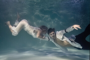 fashionable man in a white shirt and a woman in a white dress underwater in the pool