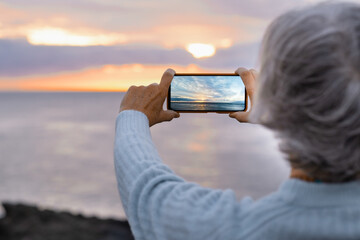 Woman holding her mobile phone taking a picture of the sunset over the sea, dramatic sky and sunlight