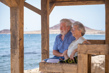 Beautiful caucasian senior couple sitting in the shade of the gazebo in front of the sea looking at...