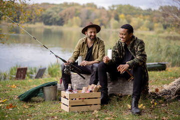 Two male friends sitting together, drinking some alcohol from flasks while fishing near lake. Caucasian and latin man at picnic outdoors. Concept of male friendship and alcohol drinks