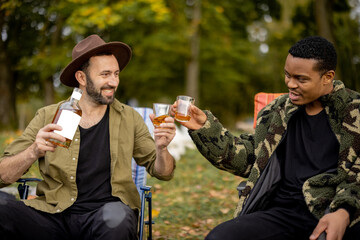 Naklejka premium Two guys having fun, drinking alcohol while fishing together on the lake coast. Caucasian and latin man resting on nature. Concept of male friendship and alcoholic drinks