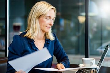 Smiling businesswoman with documents looking at laptop