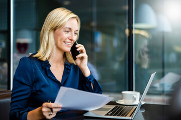 Happy businesswoman talking on smart phone and looking at laptop in cafe