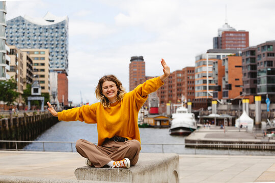 Happy young woman with arms outstretched sitting on bench at Elbphilharmonie