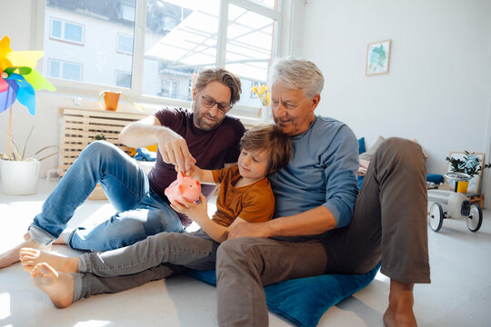 Senior Man With Son And Grandson Putting Money In Piggy Bank At Home