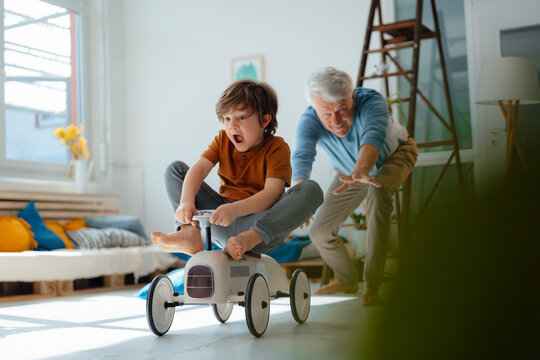 Cheerful grandfather playing with grandson sitting on toy car in living room