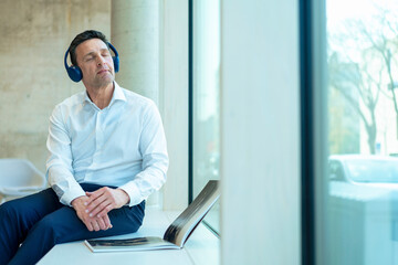 Businessman listening music on headphones sitting with magazine near window at office