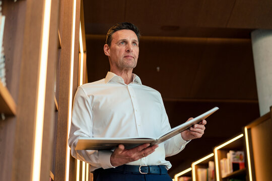 Contemplative man holding magazine leaning on rack in library