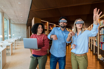 Man and woman with VR glasses gesturing by colleague holding laptop in library