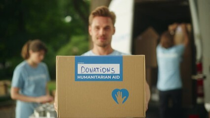 Portrait of a Handsome Young Male Volunteer. Successful Caucasian Man in Blue T-Shirt Smiling, Reaching Out with Cardboard Box to Viewer. Humanitarian Aid, Donations Center and Volunteering Concept.