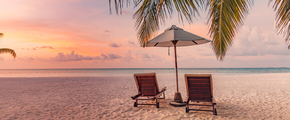 Beautiful tropical island sunset, couple sun beds chairs umbrella under palm tree leaves. White sand sea view horizon idyllic evening sky. Inspire beach resort panorama. Romantic paradise beach banner
