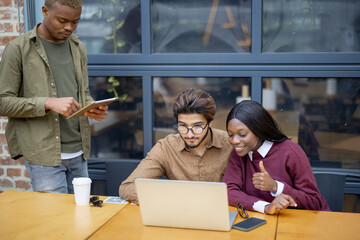 Multiracial students having video call on laptop at table outdoors. Concept of remote and e-learning. Idea of students lifestyle. Smiling young indian man, black girl and guy. University campus