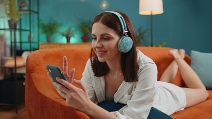 Young woman in wireless headphones relaxing lying on sofa at home choosing listening favorite energetic disco rock n roll dancing music in smartphone. People weekend activities. Happy overjoyed girl