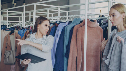Young businesswoman is checking clothes in her clothing store with tablet in hands. Her employee is...