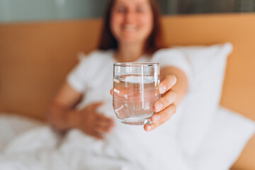 Pregnant woman with glass of water sitting on bed at home. Millennial Caucasian woman wake up in cozy white bed in modern apartment in the morning. Concept of healthy lifestyle, selective focus