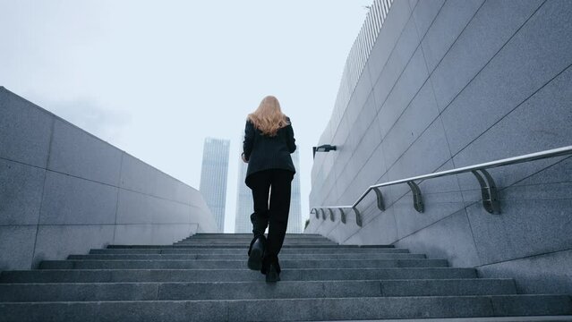 businesswoman is going to office in city center, walking up on stairs outdoors, rear view