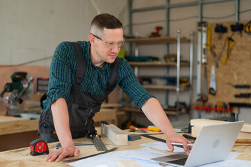 Portrait of a carpenter in protective glasses and work overalls uses a laptop in a workshop.