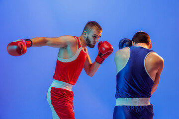 Fototapeta na wymiar Right hook. Male professional boxers in red and blue sports uniform boxing isolated on blue background in neon light. Sport, skills, power, training, energy