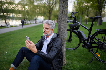 Fototapeta premium Businessman with bike sitting on grass in park, using smartphone. Commuting and alternative transport concept