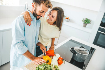 Young happy couple preparing healthy meal in kitchen at home - Husband and wife cooking salad - Food and healthy lifestyle concept - obrazy, fototapety, plakaty