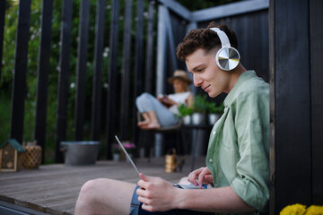 Happy young couple with laptop and headphones resting outdoors in a tiny house, weekend away and remote office concept.