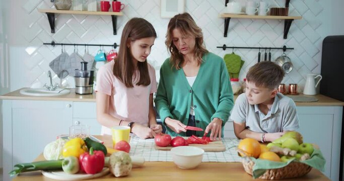 Caucasian mother teaching her younger children cutting fresh vegetables cooking salad staying together in kitchen. Home family.