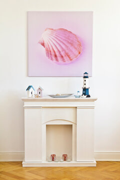 Interior shot of a square canvas print of a pink shell picture and a faux fireplace, against the white wall of an old building with parquet flooring.