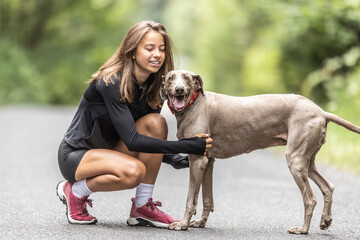Teenage girl knees and pets her Weimaraner dog during a walk in the nature