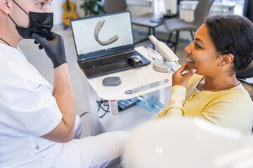 Male doctor and girl patient are sitting near laptop and talking about teeth whitening - obrazy, fototapety, plakaty