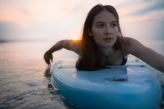 Young beautiful girl surfer paddling on surfboard on the lake at sunrise