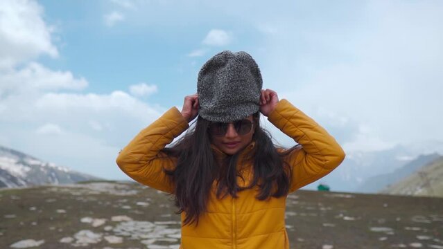 Portrait of young Indian female wearing winter jacket and warm cap while standing against snow covered Himalayan mountains at Rohtang Pass, Manali, India. Girl wearing sunglasses. Winter in Manali