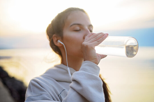Young beautiful sportive girl listening to music and drinking water at sunrise by the lake.
