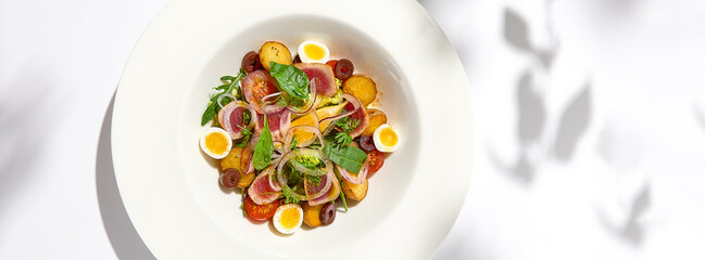 Traditional french salad nicoise with tuna, potatoes, eggs and vegetables on white table with harsh shadows. Tuna salad with quail eggs on white background with shadows of leaves.