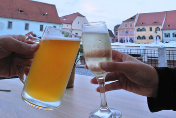 A mug of beer and a glass of champagne touch each other, to toast, at the terrace of a restaurant. Colored buildings stand out in the blurred background.