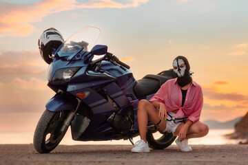 Obraz premium Independent woman in a black balaclava with skull poses near a tourist motorcycle. Sunset in the background. The concept of moto travel and motorcycle world day