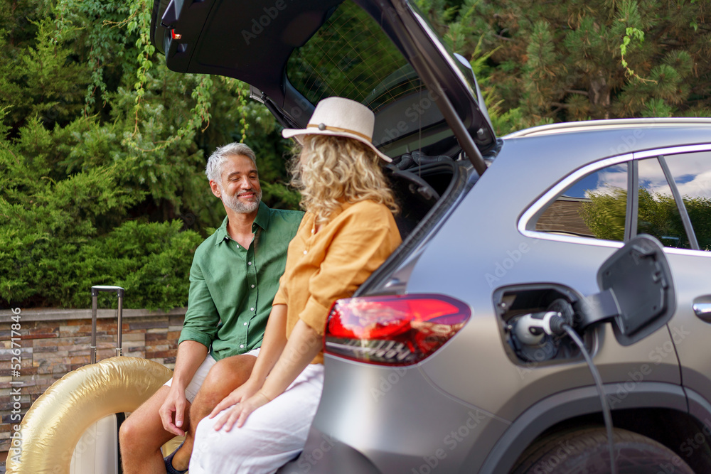 Wall mural middle-aged couple sitting in trunk while waiting for charging car before travelling on summer holid