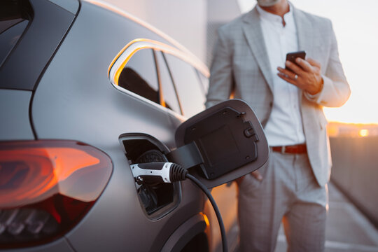 Man Holding Smartphone While Charging Car At Electric Vehicle Charging Station, Closeup.