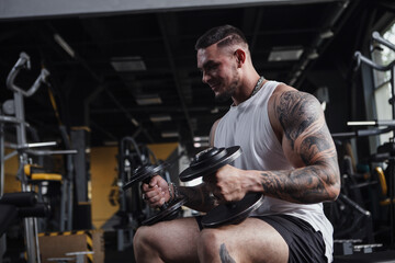 Fototapeta na wymiar Handsome tattooed muscular bodybuilder holding heavy dumbbells, sitting at the gym