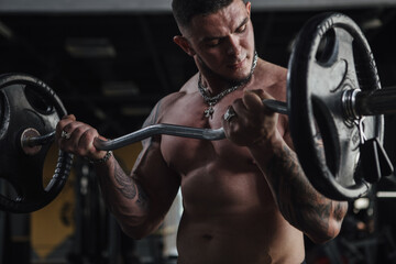 Hndsome tattooed muscular sportsman exercising shirtless at gym, doing barbell biceps curls - obrazy, fototapety, plakaty