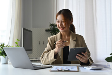 Cheerful smiling focused Asian businesswoman working on laptop holding document paper for preparing report and analyzing data resultsat at workplace.