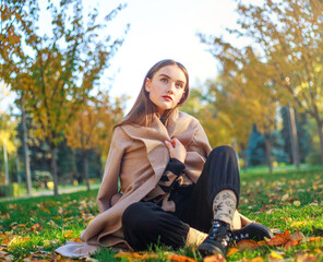 Girl in beige coat on background of autumn park and yellowed leaves on tree, thoughtfully sits on green meadow, fashion portrait young woman