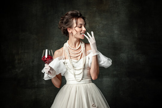 Young charming girl in image of medieval renaissance person or viscountess tasting wine isolated on dark vintage background. Comparison of eras