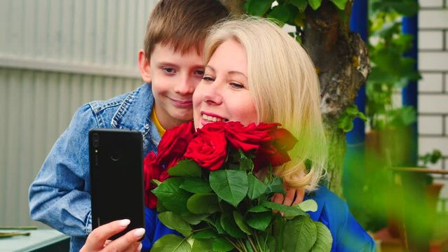 Joyful caucasian mom 45 years old holding flowers with her son 8 years old posing in front of a smartphone camera on a summer day. The child gives a bouquet of flowers to mom and is photographed
