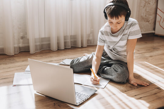 Teenager boy in wireless headphones lying on floor with laptop, having video call and online lesson at home. Schoolboy studying homework. Distance education, remote training concept.