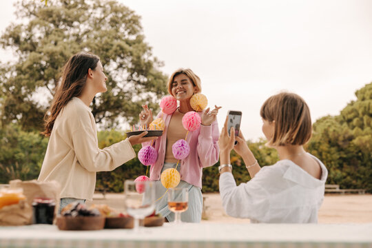 Young caucasian girl takes picture on phone of her friends celebrating birthday in fresh air. Ladies wear casual clothes outside. Leisure, technology concept