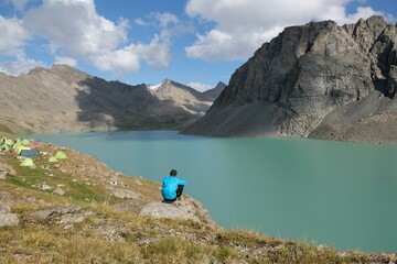 Silhouette of man sitting over lake Ala-Köl (Ala-kol, Ala-kul) in Tien Shan mountains in west of Kyrgyzstan, Asia. Cyan color of lake under steep mountaines. Touristic destination for trekking.