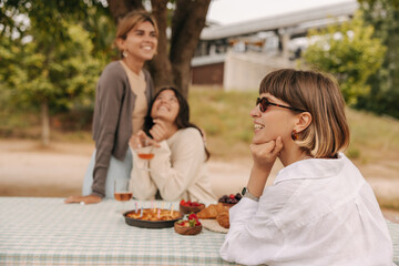 Nice young caucasian girls relax sitting at picnic table together on weekend. Women smile, wear casual clothes in spring. Nature, vacation concept