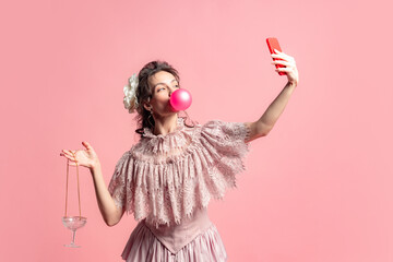 Taking selfie. One young fashionable woman with retro style hairdo wearing medieval dress posing isolated on pink background. Eras comparison concept