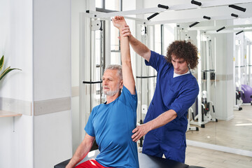 Young male doctor osteopath massaging senior man patient sitting at special bench