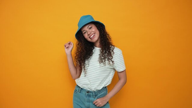 Positive brunette curly haired woman wearing panama hat showing goodbye gesture at the camera in the yellow studio